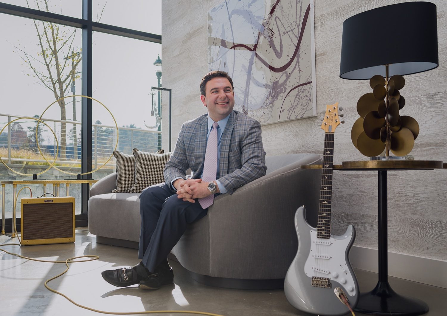 Chris Itteilag, #1 SERHANT. Agent in Washington DC and CEO & Principal of the Chris Itteilag Group, seated on a grey sofa in a modern interior beside an electric guitar and amplifier